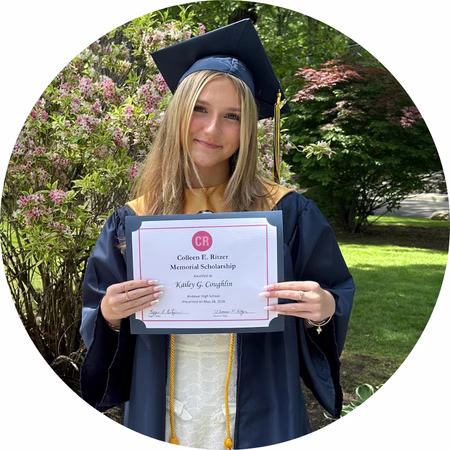 A young woman in a graduation cap and gown stands outdoors holding a scholarship certificate and smiling at the camera. Trees and greenery are visible in the background.