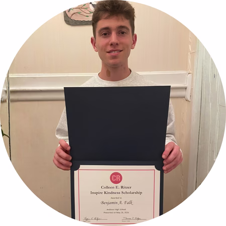 A young man stands indoors holding an open folder displaying a certificate for the Colleen R. Ritzer Inspire Kindness Scholarship. He is smiling slightly and wearing a light-colored sweatshirt.