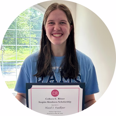 A smiling young woman with long brown hair holds a scholarship certificate in front of a sunny window, wearing a light blue T-shirt.