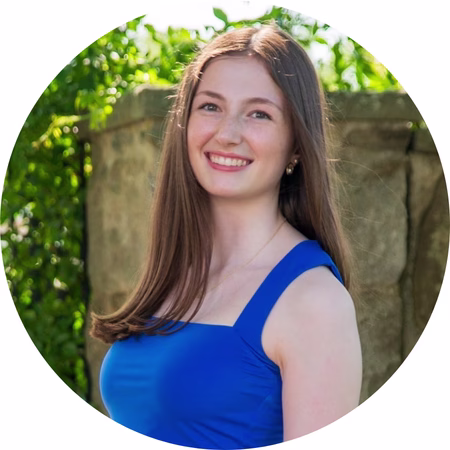 A young woman with long brown hair and a blue sleeveless dress smiles outdoors in front of a stone wall and greenery, in a circular-cropped photo.