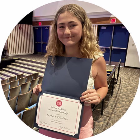 A young woman with wavy hair smiles while holding an open certificate folder in an auditorium. The certificate inside reads Colleen E. Ritzer Memorial Scholarship.