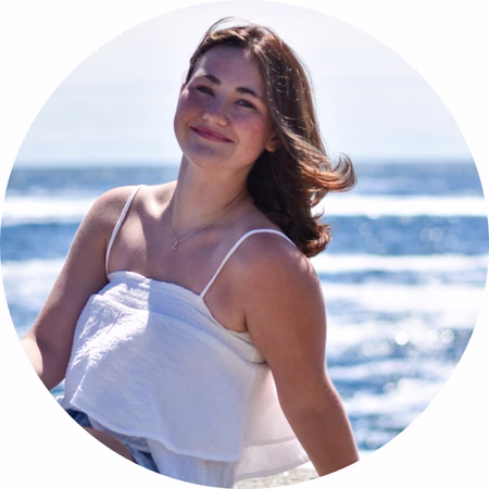 A young woman with brown hair, wearing a white sleeveless top, smiles while sitting by the ocean on a sunny day. The blue sea and sky are visible in the background.