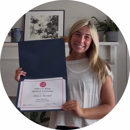 A young woman with long blonde hair smiles while holding an open folder displaying a scholarship certificate. She stands in front of a white fireplace with framed art and a plant on the mantel.