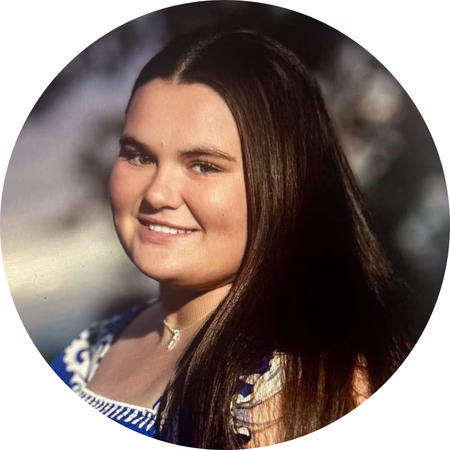 A young woman with long, straight brown hair smiles at the camera. She is wearing a blue and white patterned top and a necklace. The background is softly blurred with natural lighting.
