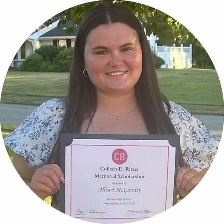 A young woman with long brown hair smiles and holds a framed certificate for the Colleen E. Ritzer Memorial Scholarship outdoors on a sunny day, with green grass and trees in the background.