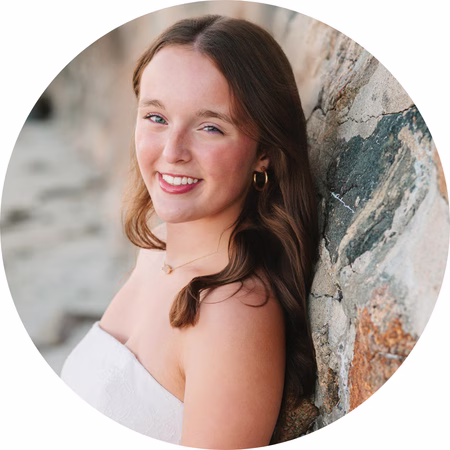 A young woman with long brown hair smiles while leaning against a stone wall. She is wearing a strapless white top and gold hoop earrings, with a blurred outdoor background.