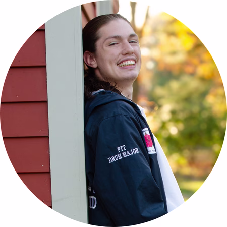 A young person with long hair and a mustache smiles while leaning against a red building outdoors. They wear a dark jacket with PIT DRUM MAJOR on the sleeve. The background is blurred with autumn colors.