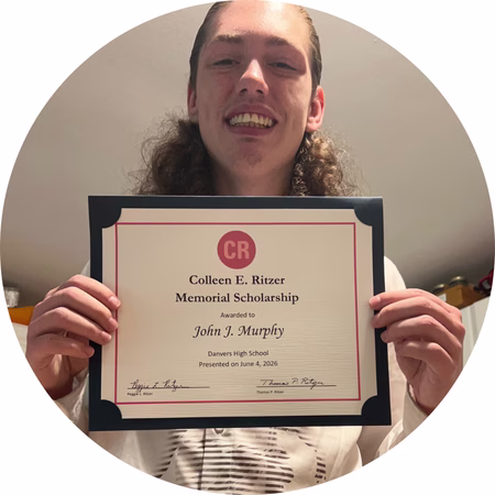A smiling person with long hair holds up a certificate for the Colleen E. Ritzer Memorial Scholarship awarded to John J. Murphy from Danvers High School.