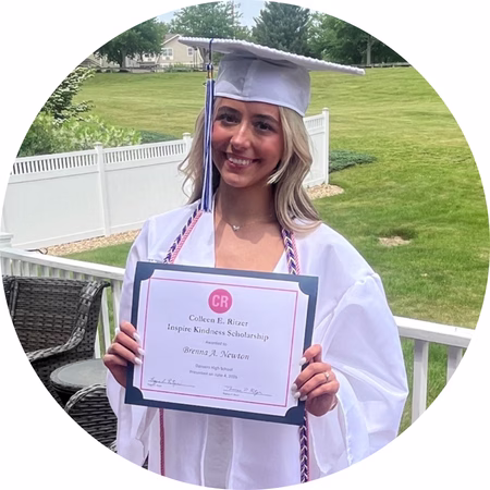 A young woman in a white graduation cap and gown smiles while holding a scholarship certificate outside on a porch, with green grass and trees in the background.
