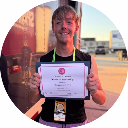 A young person standing outdoors smiles while holding a “Colleen E. Ritzer Memorial Scholarship” certificate. They are wearing a black sleeveless shirt and an event lanyard.