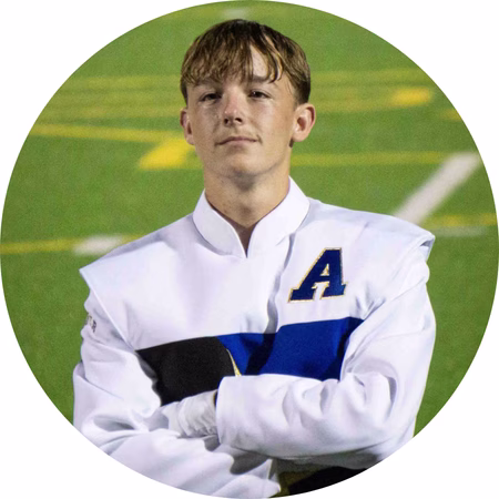 A young person in a white band uniform with a blue stripe and a letter A stands on a sports field, holding a black hat and looking confidently ahead.