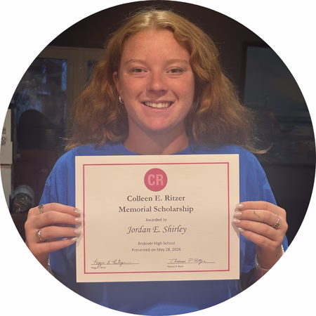 A young woman with long, wavy hair smiles and holds up a Colleen E. Ritzer Memorial Scholarship certificate awarded to Jordan E. Shirley from Andover High School.