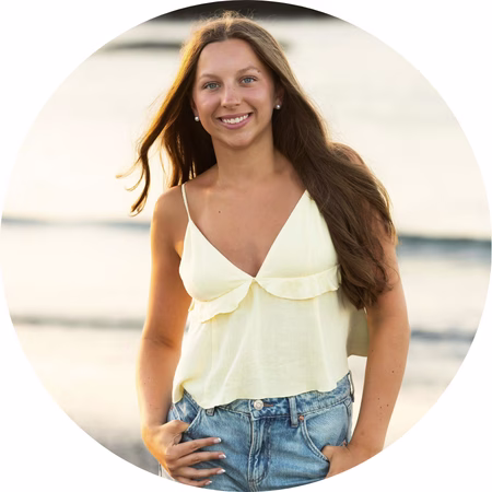 A young woman with long brown hair wearing a light yellow sleeveless top and blue jeans stands outdoors, smiling with one hand in her pocket, against a blurred background.