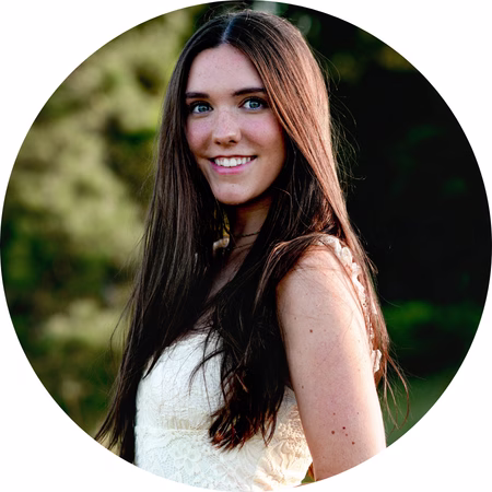 A young woman with long brown hair and blue eyes, wearing a white sleeveless dress, smiles while standing outdoors with trees in the background. The image is framed in a circular crop.