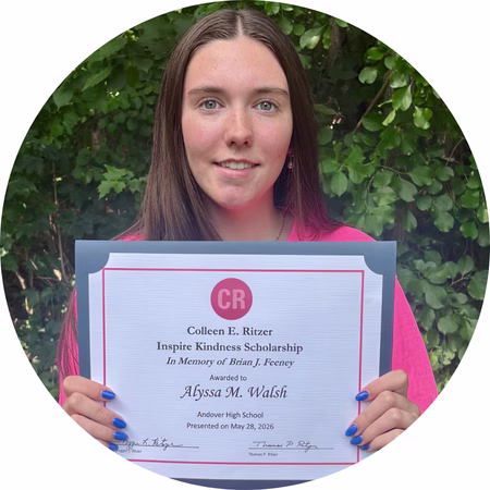 A young woman with long brown hair holds a scholarship certificate in front of green foliage, smiling slightly. The certificate is awarded to Alyssa M. Walsh from Andover High School for the Inspire Kindness Scholarship.