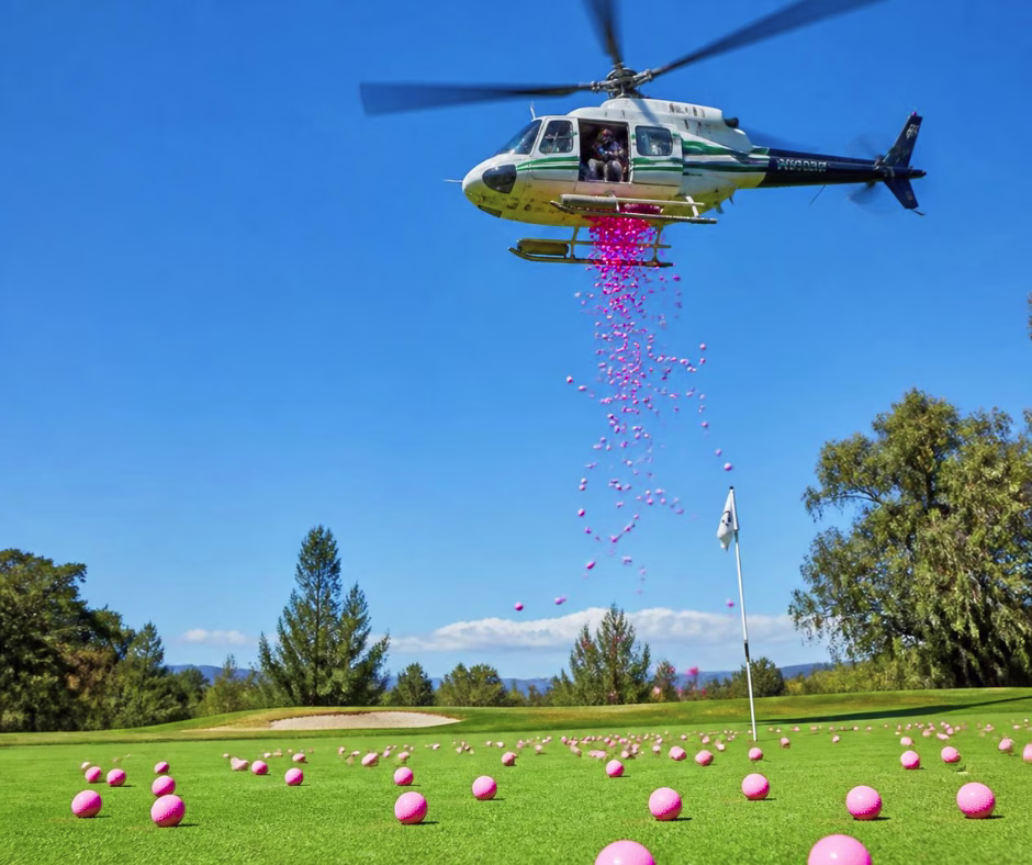 A helicopter hovers over a golf course, dropping many pink golf balls onto the green, with more pink balls already scattered around the flag.