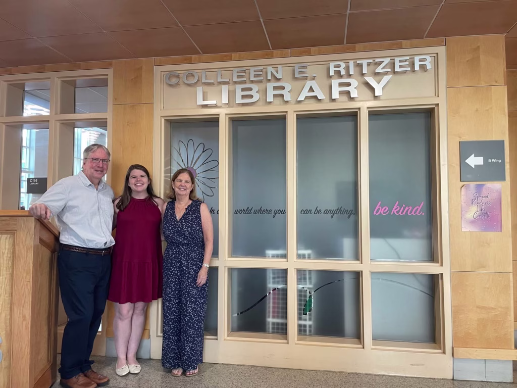 Three people stand smiling in front of the Colleen E. Ritzer Library entrance. The background features glass panels with positive messages and wooden framing.